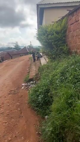 The video pans across a rural African village. The foreground features a dirt road with scattered litter and lush green vegetation. To the right, a brick wall runs alongside a building, overgrown with a large bush. In the middle ground, several people are gathered on a concrete sidewalk next to a building with columns. Further down the dirt road, more buildings with corrugated metal roofs are visible, and several people are seen near the entrances of these structures. The background reveals a hilly landscape under a cloudy sky. The overall impression is of a modest, everyday scene in a developing community.