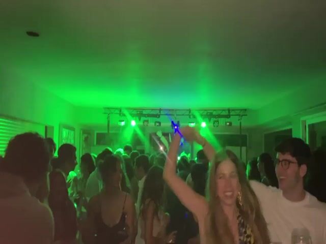 A crowd of people are dancing in a room with flashing green, red, and blue lights. Some people are holding drinks. A stage with lights is visible in the background. The camera is moving throughout the video.