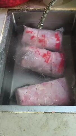 A sink filled with water contains three vacuum-sealed packages of raw meat. A faucet above the sink is spraying water onto the meat, causing bubbles and splashes. The meat packages are white with red lettering. The sink is made of stainless steel and appears to be in a kitchen or food preparation area.