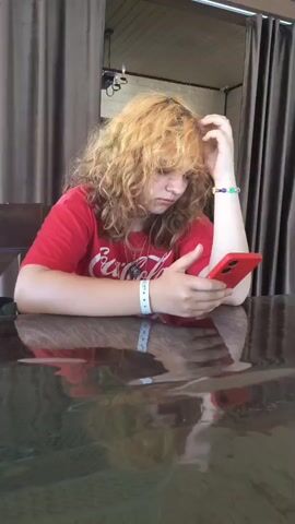 A young person with wavy blonde hair, wearing a red t-shirt, sits at a table with a reflective surface, holding a red smartphone and looking at it with a pensive expression. They scratch their head with their left hand. A bracelet with colored beads is visible on their right wrist, as is a white band on their left wrist. In the background, brown curtains hang, and the environment appears to be outdoors or on a covered patio, with a wooden ceiling visible. A menu, partially obscured and reflecting the surrounding environment, lies on the table in the foreground.
