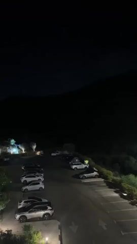 The video shows a dimly lit outdoor parking lot at night. Several cars are parked in the lot. A dark mountain or hill looms in the background. There are some lights illuminating parts of the parking lot and some vegetation. The sky is dark, indicating nighttime. The video appears to be filmed from a slightly elevated position.