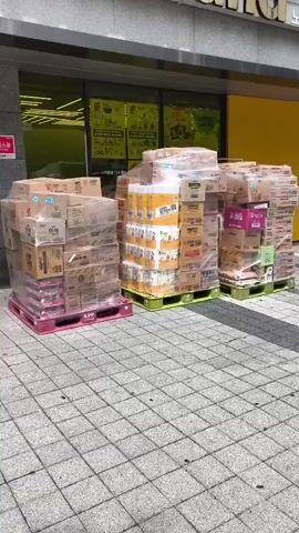 The video shows the exterior of a No Brand store. Outside the store, there are three large stacks of cardboard boxes on pallets, shrink-wrapped for protection. The boxes appear to be merchandise. The store has a glass storefront with yellow signage inside. The building facade is gray stone. The ground in front of the store is paved with interlocking gray bricks. The camera pans slightly upwards to reveal more of the building's facade and some greenery at the top. There are no people visible in the frame.