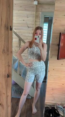 A young woman with long auburn hair is taking a mirror selfie in a room with wood-paneled walls. She is wearing a matching leopard-print workout set consisting of a zip-front crop top and high-waisted bike shorts. She is standing on a wooden floor, and her reflection shows a staircase to her left, a doorway to her right, and a shelf with shoes in the background. She has her left hand on her hip and is holding her phone in her right hand. The woman is smiling and appears to be posing for the photo.