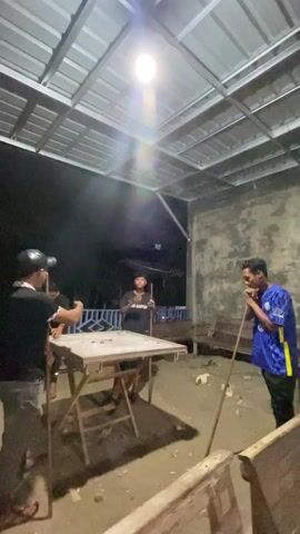 The video shows four young men playing carrom in an outdoor, covered area at night, likely in Khang Tanjong, Indonesia. The subjects are four males, appearing to be in their late teens or early twenties. One person, wearing a black t-shirt and a black baseball cap, is positioned to the left of the carrom table, holding a striker. Another person, wearing a black t-shirt, stands on the opposite side of the table, also holding a striker. The third person, wearing a blue shirt with yellow accents and black pants, stands to the right of the table, holding a striker. The fourth person, wearing a black t-shirt, is visible behind the carrom table.

The primary object is a carrom board, set up on a folding table. The playing surface of the carrom board is visible, with several coins scattered on it. Other visible objects include the strikers, which are disc-shaped objects used to hit the carrom coins, and wooden benches in the background.

The scene is set in a covered outdoor area, possibly a patio or an extension of a building. The roof is made of corrugated metal sheets, and a bright light bulb hangs from the ceiling, illuminating the playing area. The background suggests a rural or semi-rural setting with darkness beyond the illuminated space. The ground appears to be dirt or sand.

The activity taking place is a game of carrom. The men are holding strikers and seem to be in the process of playing, with at least two of them actively engaged with the game. The mood appears casual and social. The time of day is clearly night, indicated by the darkness in the background and the artificial lighting. No text is visible in the video.