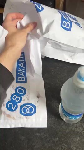 A person's hand opens a white plastic bag with a blue "BAKARI" logo. Inside the bag are pastries, including brown rolls. There is a red smear on the inside of the bag. In the background, there are other similar bags and a bottle of water. The countertop is dark and speckled.