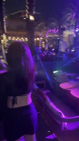 The video shows a woman in a black crop top and skirt walking through an outdoor nightlife venue. There are palm trees and string lights. People are seated and walking around in the background. The lighting shifts between purple and blue.