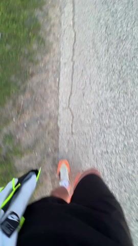 The video opens with a first-person view, looking down at a person's feet as they walk or run on an asphalt path next to a grassy area. The person is wearing black shorts and orange and white running shoes. A light grey bag with a neon green strap is visible on their left side.

The camera then pans up to reveal a stunning sunset over a rural landscape in Bohot, Bulgaria. The sky transitions from deep blue at the top to vibrant orange and yellow hues near the horizon, where the sun is setting. A paved road stretches into the distance, flanked by green fields and some darker, possibly tilled, land. Small trees and bushes dot the scenery. The overall atmosphere is peaceful and serene at dusk.