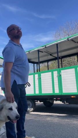 The video shows a man with glasses and a blue shirt petting a large white dog next to a small green and white tour train on a sunny day. The man looks up at the sky. Several people walk behind the man and dog and toward the train. The sky is blue with some white clouds. The tour train has several carriages behind the engine.