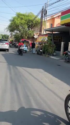 The video shows a street scene with several motorcycles and a cart being pulled by a motorcycle. The camera is mounted on a motorcycle and is looking forward, showing a person in the foreground wearing a blue t-shirt and shorts, with their legs visible. In the background, there are buildings, trees, and other motorcycles on the road. The overall impression is a busy street in a city or town.