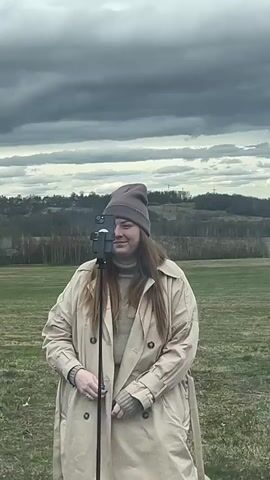 A woman stands in a field with her hands folded at her waist and wearing a tan coat and matching beanie. The woman adjusts the phone in front of her, which is mounted on a tripod. In the background is a green field, rolling hills and a line of trees. The sky is dark and cloudy.