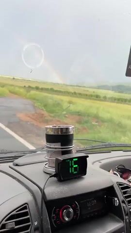 The video shows a view from inside a car driving on a wet road. A rainbow is visible in the distance, spanning over green fields and rolling hills under a cloudy sky. Raindrops are visible on the windshield. The car’s speedometer displays a reading of 76 km/h. An air freshener hangs from the rearview mirror. An ashtray is visible on the dashboard.