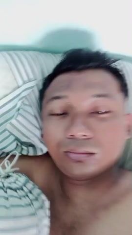 A bare-chested man with short black hair and brown skin is lying in bed, positioned on a striped pillow and partly covered with a striped blanket. His gaze is mostly directed at the camera, although he occasionally looks down. The background consists of a plain, light blue wall. The camera angle is a close-up, personal perspective.