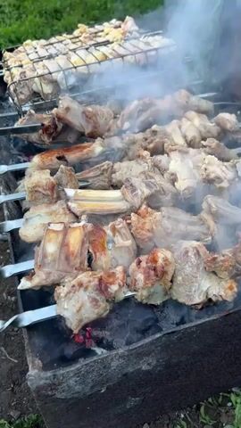 Meat is cooking on a grill. There are many pieces of meat on several skewers. Some pieces are ribs. Some pieces are cuboid. The meat is mostly brown and grey. There is white smoke coming from the meat and grill. In the background, there is a grilling basket on a grill with white meat. The ground is green and covered in grass.