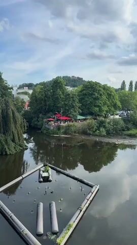 The video shows a wide shot of a calm river with a floating structure in the foreground. The riverbank on the opposite side is lined with lush green trees and vegetation. A small outdoor cafe with red umbrellas is visible nestled amongst the trees, with patrons seated at tables. In the background, a town or city skyline is visible, with buildings and a hill topped with a structure, possibly a castle or tower. The sky is partly cloudy, with a mix of blue sky and white clouds. The overall scene depicts a tranquil, scenic view of a riverside town on a pleasant day.
