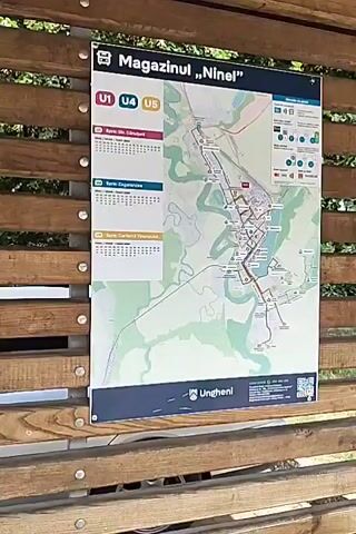 The video begins at a bus stop with a map displayed on a wooden structure. The map shows bus routes labeled U1, U4, and U5. The bus stop appears to be located in a town or city. The camera then pans to the right, revealing a park-like area with trees, benches, and a trash can. A sidewalk made of paving stones is visible, along with a street and a pedestrian crossing sign. The video ends with a view of the surrounding area, including buildings and more trees.