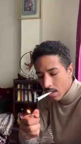 A man with curly hair and a mustache is lighting a cigarette with a lighter. He is wearing a tan turtleneck sweater. He takes a drag and exhales a large cloud of smoke toward the camera. He looks into the camera while exhaling. Behind him is a wooden cabinet with books and various small objects on it. A framed painting hangs on the wall above the cabinet. A maroon curtain is visible on the right.