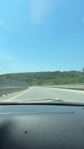 The video shows a view from inside a car driving on a highway. The sky is clear and blue. On either side of the highway are barriers and green hills covered in trees. The dashboard of the car is visible in the foreground.