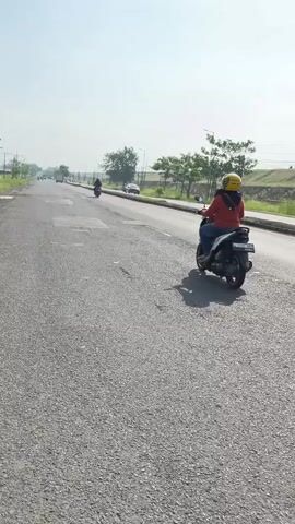 A person wearing a yellow helmet and an orange jacket rides a black scooter on a paved road. The road has patches of damage. In the distance, several other vehicles, including another scooter and cars, are visible. The environment appears to be outdoors, with trees and grassy areas lining the side of the road. The sky is bright and clear.
