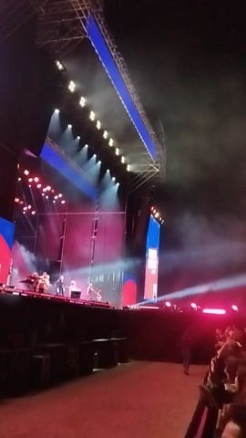 The video shows an outdoor nighttime concert or live music event taking place in Maipú, Chile. The scene is dominated by a large stage, brightly lit with numerous spotlights and colored lights, primarily red and blue. Several performers, appearing to be a band, are on stage. One musician is prominently visible on the left side of the stage, standing behind a keyboard or synthesizer. Other band members are situated further back on the stage.

Large LED screens form the backdrop and sides of the stage, displaying dynamic visual effects. On the left screen, partial text "S AS" is visible, accompanied by snowflake graphics, suggesting a holiday or Christmas-themed event. Another screen on the right shows a large red circular graphic with blue and white elements. Smoke or fog effects are emanating from the stage, enhancing the theatrical atmosphere. The overall setting indicates a large-scale, energetic live performance.