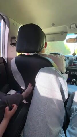 The video shows the interior of a car from the perspective of a passenger in the back seat. The driver, a person with short hair, is visible in the front seat, gripping the steering wheel. Two other passengers are also in the back seat, with their legs visible in the foreground. One passenger has their bare feet resting on the seat. The car appears to be moving down a road lined with trees. The sun is shining, casting light into the car. A phone is mounted on the dashboard.