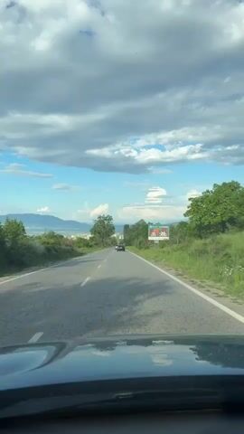 A car drives down a road toward a city in the distance. The road is lined with trees and shrubs. A billboard is seen on the right. The sky is blue with scattered clouds. Another car is slightly ahead. The camera is mounted inside a car behind the windshield. Someone speaks.