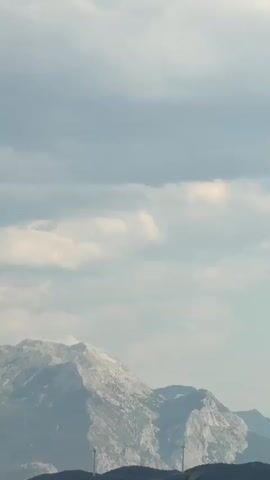 The video shows a wide shot of a mountain range under a cloudy sky. The mountains are rocky and appear to have some snow or light-colored patches on their peaks. In the foreground, there are dark green trees. There are also a few wind turbines visible on the distant hills. The overall impression is of a natural landscape with some man-made structures.