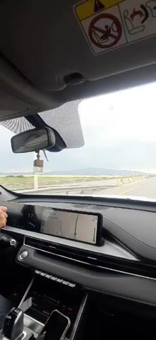 The video shows the interior of a car driving on a highway. The dashboard is visible, with a navigation screen displaying a map. The windshield shows a road stretching into the distance, with yellow and white lines marking the lanes. On the right side of the car, a side mirror reflects the passing scenery, which includes rolling hills and a cloudy sky. There are no people clearly visible inside the car. The car appears to be moving at a steady speed on a clear day.