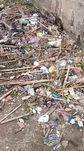 A video shows a pile of garbage and debris in an outdoor area. The garbage includes plastic bags, sticks, leaves, and what appear to be shoes and other discarded items. The area appears to be a drainage ditch or channel, with a concrete wall on one side. There is a small amount of murky water visible in the channel. The video pans across the scene, showing the extent of the garbage accumulation.