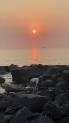 A young boy walks across dark-colored rocks at the edge of the sea. The sun is setting over the water in the background, creating a hazy orange sky. A boat is faintly visible in the distance.