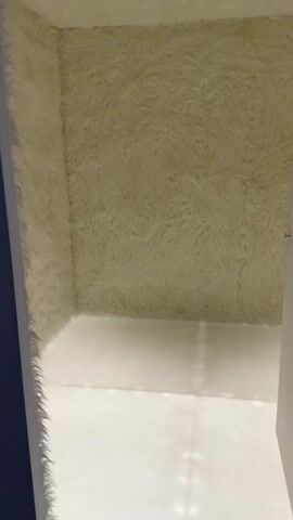 The video begins by showing the corner of a room where the walls are covered in a shaggy, cream-colored material. The floor appears to be a white, possibly tiled surface, reflecting light. The camera pans across the furry walls, revealing their texture and depth. As the video progresses, the perspective shifts to a top-down view, focusing on a plush, white, shaggy rug. A hand with rings on its fingers reaches into the frame, holding a black camcorder. The hand then moves the camcorder across the surface of the rug, touching the fibers. The video appears to be a close-up shot of the rug and a hand interacting with it while holding a camcorder.