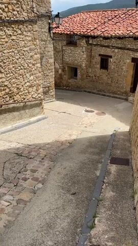 The video shows a narrow, sloped street in a village. The buildings on either side are made of stone with red tile roofs. The sky is partly cloudy with mountains in the background. The street is paved with a mix of concrete and cobblestones. There are shadows cast by the buildings, indicating a sunny day. No people or animals are visible.