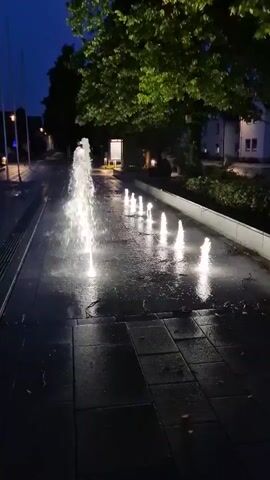 The video shows a row of illuminated fountains spraying water upwards in a dark, outdoor setting. The fountains are arranged in a line, with one larger fountain in the foreground and several smaller ones receding into the background. The water from the fountains is lit from below, creating a glowing effect. The ground around the fountains is wet and reflective, indicating that the water is splashing onto the pavement. In the background, there are trees and buildings, as well as streetlights. The overall impression is one of a serene and tranquil evening scene.