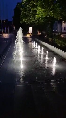 Illuminated fountains glow across a paved square in Ergolding at dusk