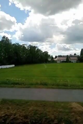 The video shows a view from a moving vehicle. The scene depicts a rural landscape with green fields, trees, and a cloudy sky. There are several white wrapped bales of hay or silage lined up in a field. In the distance, there are some buildings, possibly houses. The road or path in the foreground is visible. The video captures the dynamic scenery as the vehicle moves through the countryside.