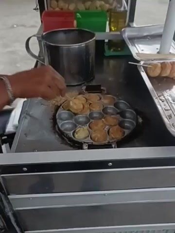 The video shows a person preparing food on a street cart in Keude Lueng Putu, Indonesia. The person, wearing a silver bracelet on their left wrist, is seen using wooden skewers to flip small, round, golden-brown food items being cooked in a specialized circular pan with multiple molds. These food items resemble small meatballs or dumplings. To the right of the cooking pan, a metal tray holds several skewers of the cooked food, indicating they are ready for serving.

The street cart has a metal cooking surface and appears to be set up for mobile food vending. Behind the cooking area, there are containers of ingredients, including a red container, a green container, and a bottle of oil. Above these, a glass display case shows more of the cooked food items. Visible text on the cart reads "MEULABOH".

The scene suggests a daytime activity, possibly a market or a busy street, given the presence of the cart and the implied outdoor setting. The focus of the video is on the food preparation process, highlighting the meticulous work of the vendor. The food items being cooked appear appealing, and the overall impression is one of a lively street food vendor in operation. The lighting suggests it is daytime, but there are no strong indicators of specific weather conditions.