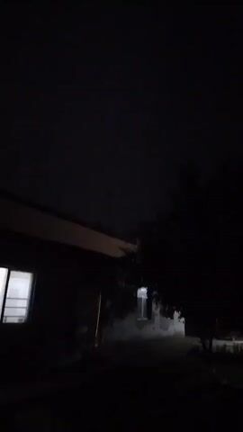 A blurry, dark video captures the exterior of a building at night. The sky is black, suggesting darkness or heavy cloud cover. A portion of a building's roofline is visible on the left, with a lit window peeking out from its side. To the right, a dark tree stands silhouetted against the faint light of another window. The ground is indistinct, and the overall scene is obscured by low light and motion blur.