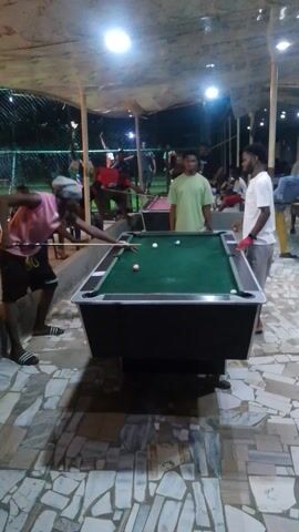 The scene appears to be an outdoor recreation area with a pool table as the central focus. The video is shot from a low angle and rotated. There are several individuals present, one poised to take a shot at the pool table. Other individuals are standing around observing the game. The lighting is bright with overhead lights. The ground is paved with a mosaic tile pattern. The overall atmosphere seems casual and social.