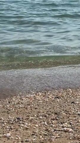 The video shows a close-up view of small waves gently washing onto a beach covered in sand and pebbles. The water is clear and reflects the sunlight, creating a shimmering effect. The waves are calm and create a soft white foam as they reach the shore. The beach appears to be undisturbed, with no people or objects visible. The overall scene is peaceful and serene, evoking a sense of tranquility and the natural beauty of the seaside.
