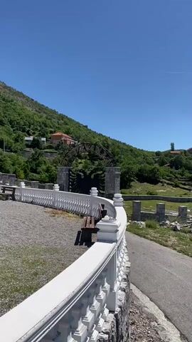 A vertical video pans across a scenic landscape under a clear blue sky. In the foreground, a white ornate railing curves along a paved path. Beyond the railing, a small park area with a wooden bench is visible. Further into the scene, a road winds upwards, flanked by green hills and scattered stone ruins. A large, dark, circular structure, possibly a water wheel or part of an old mechanism, is perched on the hillside. Several stone pillars, remnants of a structure, stand amidst piles of rubble. In the distance, houses and buildings dot the landscape, nestled among lush green trees and rolling hills. Mountains form a backdrop against the sky, with a few scattered clouds. The overall impression is one of historical ruins set within a beautiful, natural environment.