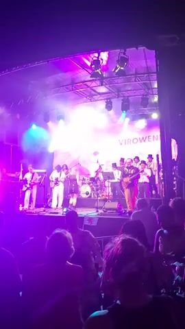 A live band performs on an outdoor stage under bright purple and white lights, with a large banner behind them displaying the text "VIROWE NATIONAL F". Several band members are visible, including individuals playing guitars, drums, and a saxophone. The audience, silhouetted in the foreground, watches the performance. The stage is equipped with multiple spotlights and speakers. The overall atmosphere appears to be a lively outdoor concert or festival.