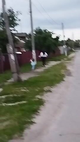 A person wearing a white shirt and black shorts is standing near a maroon fence. Next to them, a child wearing a light blue shirt and light-colored shorts is running away from them. The two are on a dirt path with grass on the left side and a road on the right. There are telephone poles with wires overhead. Houses are visible in the background. The sky is cloudy.