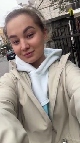 The video shows a young woman taking a selfie video while walking outdoors. She's wearing a beige jacket over a white hoodie and a light blue shirt. The setting appears to be a residential area with buildings, cars, and trees in the background. The sky is overcast. The woman looks directly at the camera, smiling and slightly turns her head.