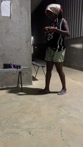 The video shows a Black woman dancing outside. She is wearing a headscarf, basketball jersey, and light-colored shorts, and dancing in flip-flops on a concrete surface. To her left is a small table with various objects on it, and behind the table are concrete walls with corrugated metal paneling. The video is lit dimly.