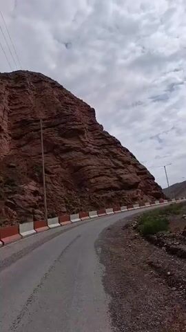 The video shows a POV shot from a vehicle driving on a paved road. The road curves to the left, with a steep, reddish-brown cliff face on the left side and a gentler, rocky slope on the right. The road is bordered by a white and orange barrier on the left. There is some sparse green vegetation growing on the right side of the road. In the distance, a tall, thin street lamp stands beside the road. The sky is cloudy, with patches of blue sky visible. Power lines are also visible strung between poles in the distance. The road appears to be in a mountainous or canyon-like area.