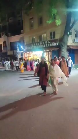 A nighttime street scene shows a large group of people walking down a street. The people are dressed in various types of clothing, including traditional Indian attire like saris. Some people are wearing white robes. The group is walking in the same direction.  On the right side of the frame is a restaurant called "SAIGOKUL REEN TREE RESTAURANT".  There are buildings lining the street.  A motorcycle is parked on the left side of the frame. Streetlights illuminate the scene. Shadows are cast on the road.