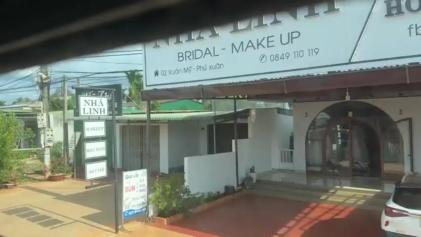 A video recording captures a view from a moving vehicle. The video opens with a shot of a building with a sign that reads "Nha Linh Studio - Make Up - Bridal - Nail." The building appears to be a business, possibly a salon or beauty parlor. The video then pans to the right, revealing more of the surrounding environment. The scene includes various buildings, fences, trees, and power lines. There are also glimpses of vehicles, including a truck and a motorcycle. The overall environment seems to be a suburban or rural area. The video continues to pan, showing more residential buildings and lush vegetation. The sky is overcast with clouds, suggesting a cloudy or potentially rainy day. The camera movement is smooth, indicating that the vehicle is traveling at a moderate speed. The video ends with a shot of a house with a gated entrance and a well-maintained garden. There are no visible people in the video.