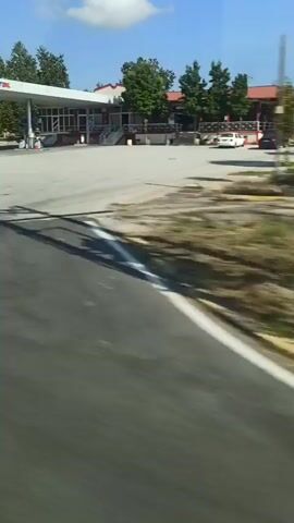 The video shows a view from a moving vehicle, likely a bus or car, traveling on a road. The camera is pointed towards the right side of the vehicle.

In the initial frames, a gas station is visible with pumps under a canopy. Trees and some buildings are also present in the background. The road has yellow and black striped barriers along its side.

As the vehicle moves, the scene transitions to a more rural landscape. Open fields with dry grass and green vegetation are visible, with rolling hills in the distance under a clear blue sky with a single white cloud. More trees line the side of the road.

Later frames show a brief glimpse of houses or buildings with red roofs amongst dense green foliage. The movement of the vehicle causes a blurring effect in all parts of the video, making detailed observation of specific objects or individuals challenging. There are no people clearly visible or identifiable in the video. The overall impression is of a journey through a mix of developed and natural environments.