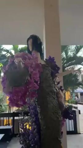 The video shows an indoor scene with a flower-covered mannequin and a fountain. The mannequin is covered in green foliage and purple orchids, with black hair. The fountain is a square pool with multiple jets of water shooting upwards. Beyond the fountain, palm trees and a glimpse of a building can be seen through an open walkway. A sign is visible next to the fountain, and the overall setting appears to be a well-lit, indoor mall or atrium.