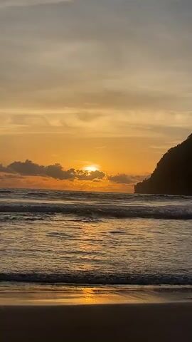 The video shows a sunset over a beach. The sun is low in the sky, casting a warm orange glow. There are clouds in the sky, and the ocean waves are gently rolling onto the shore. A dark silhouette of a hill or cliff is visible on the right side of the frame. The foreground is the sandy beach.