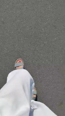 The video shows a first-person perspective of someone walking on an asphalt surface. The person is wearing light-colored pants and light-colored sandals with straps. The ground appears to be a sidewalk or path, with some cracks and texture visible in the asphalt. The lighting suggests it is daytime. The camera is pointed downwards at the person's feet and the ground as they walk.